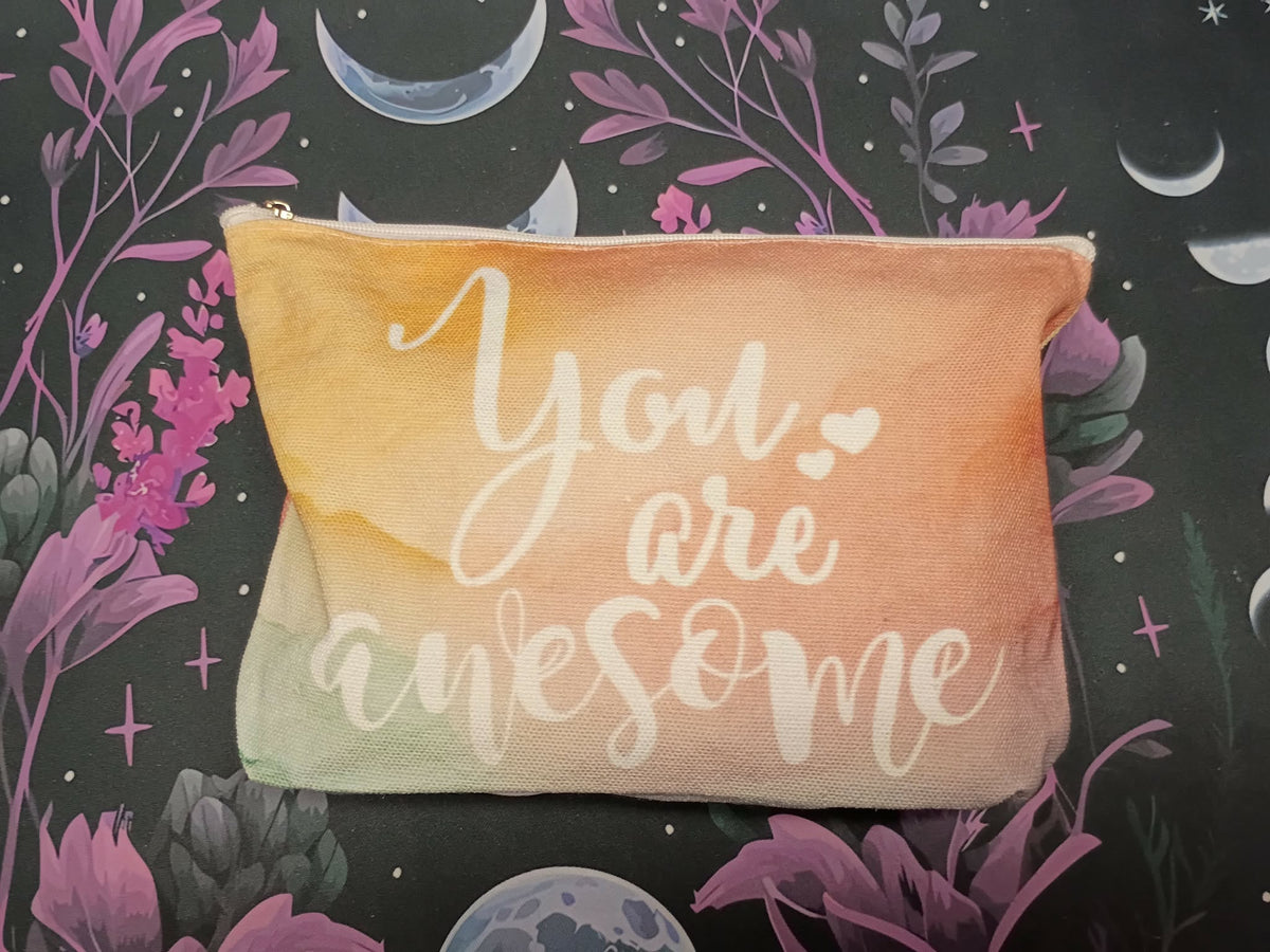 A rectangular zippered pouch with a watercolor gradient transitioning from peach to pale yellow and mint green, featuring the phrase 'You are awesome' in white script. The pouch is laid flat on a dark patterned background with purple flowers, pink blossoms, and celestial illustrations.